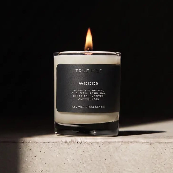 A lit True Hue 'Woods' scented candle in a clear glass jar with a black label, sitting on a textured light gray stone pedestal against a dark background.