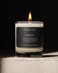 A lit True Hue 'Woods' scented candle in a clear glass jar with a black label, sitting on a textured light gray stone pedestal against a dark background.