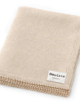 A folded beige knit blanket with a textured ribbed edge and a white label reading 'Bleu-La-Lo ORGANICS', shown on a clean white background.