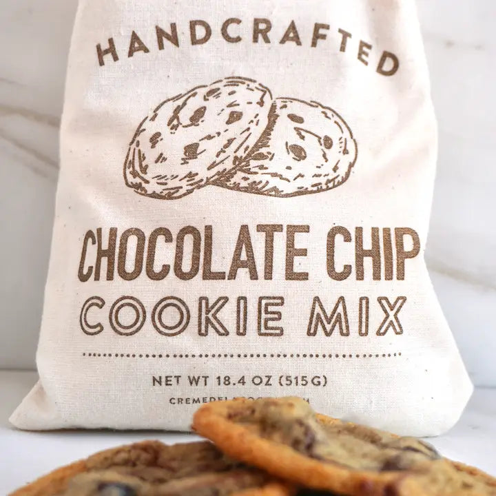A cream-colored canvas bag printed with 'HANDCRAFTED CHOCOLATE CHIP COOKIE MIX' and an illustration of cookies, with chocolate chip cookies placed in front on a light marble surface.