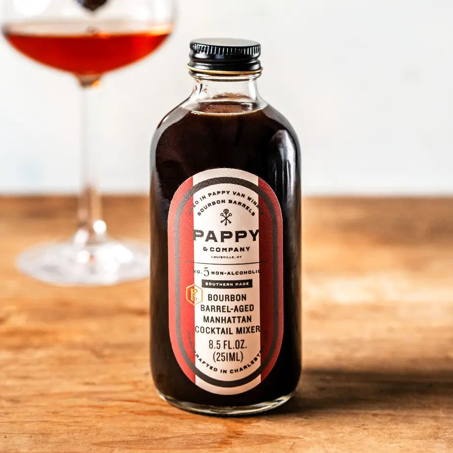A bottle of Pappy &amp; Company Bourbon Barrel-Aged Manhattan cocktail mixer sits on a wooden table with a blurred glass of the reddish-brown drink in the background.
