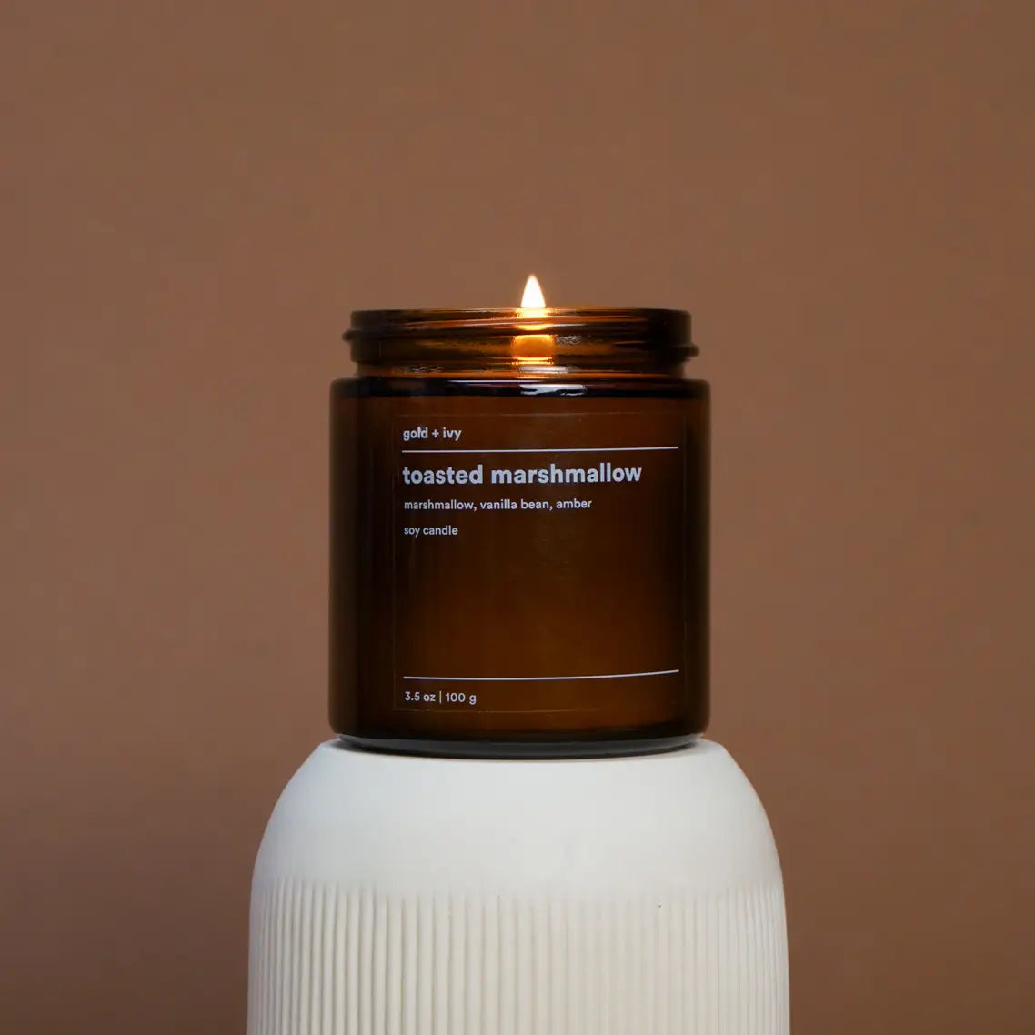 Candle in a brown jar labeled 'toasted marshmallow' on a white pedestal against a brown background