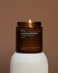 Candle in a brown jar labeled 'toasted marshmallow' on a white pedestal against a brown background
