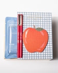 A boxed set containing a blue packet of NARS Hand Sanitizer, a red pen, and a decorative box with a blue gingham pattern and a large red apple illustration, photographed on a clean white background.