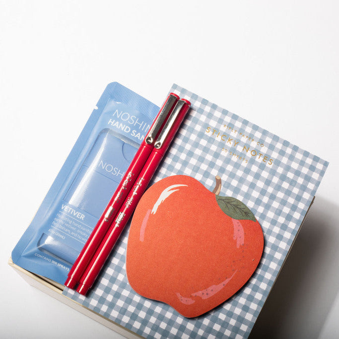 A boxed beauty gift set on a white background, featuring a blue hand cream packet, two red cosmetic pencils, and a decorative box lid with a blue gingham pattern and a red apple illustration.