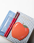 A boxed beauty gift set on a white background, featuring a blue hand cream packet, two red cosmetic pencils, and a decorative box lid with a blue gingham pattern and a red apple illustration.