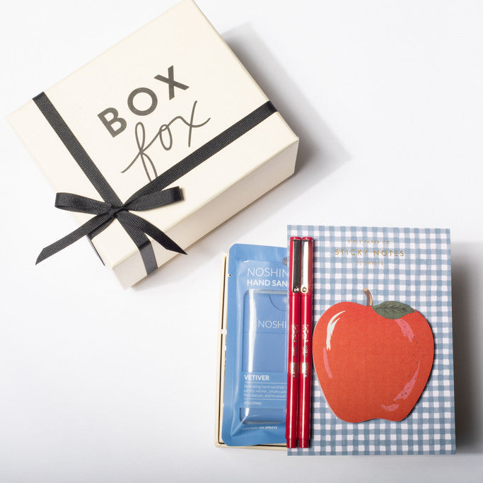A curated gift set featuring a cream-colored 'BOX fox' box with a black ribbon, alongside a blue and white gingham notebook with a red apple illustration, a blue packet, and a red pen, all arranged on a clean white background.