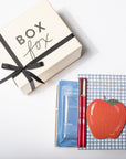 A curated gift set featuring a cream-colored 'BOX fox' box with a black ribbon, alongside a blue and white gingham notebook with a red apple illustration, a blue packet, and a red pen, all arranged on a clean white background.