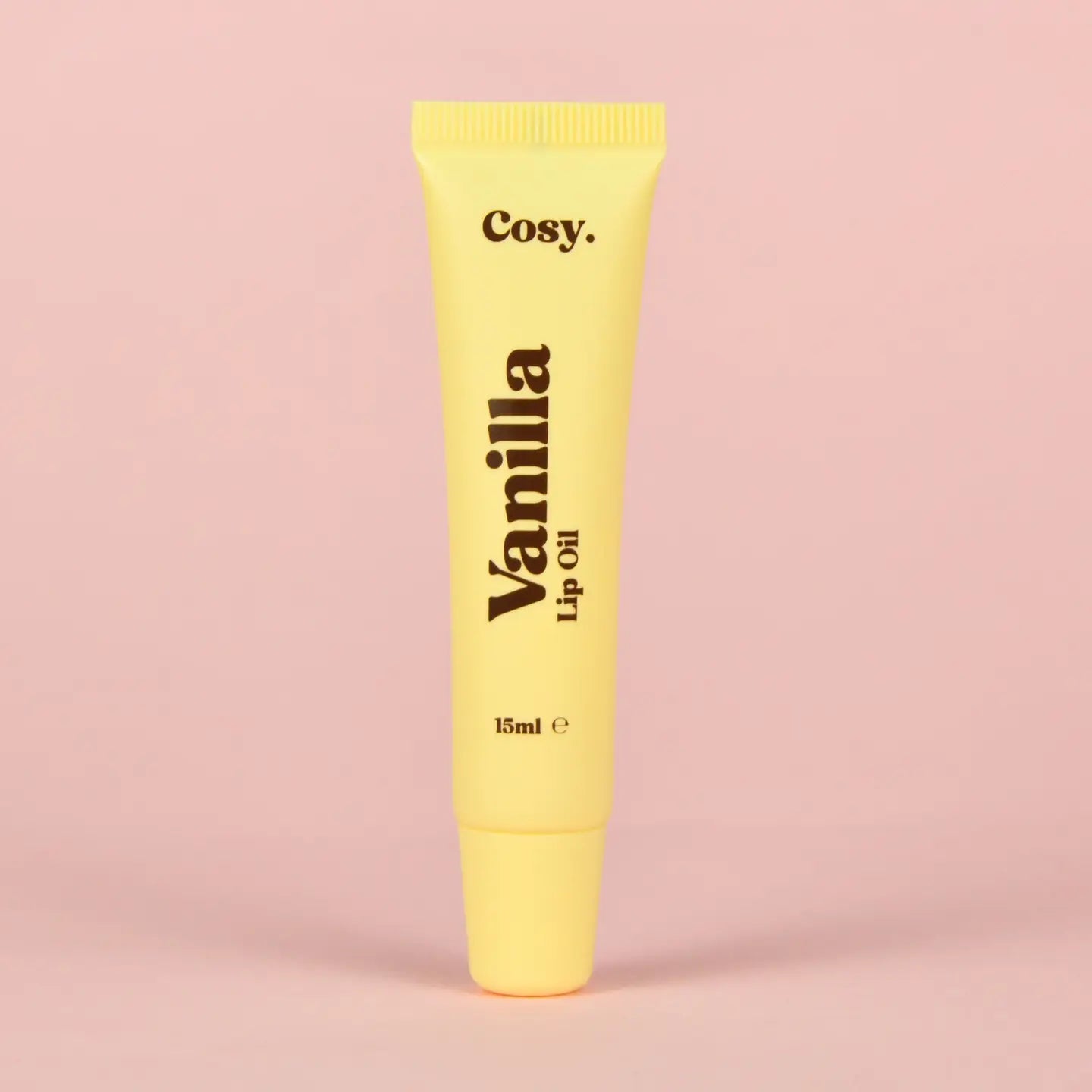 Yellow lip oil tube labeled 'Vanilla' with 'Cosy.' brand on a pink background