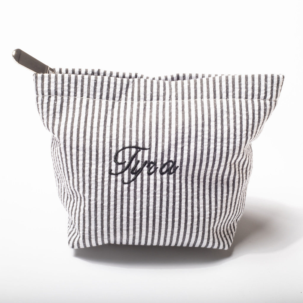 A gray and white vertically striped fabric pouch with a zipper closure and the name 'Tyra' embroidered in black script on the front, set against a plain white background.