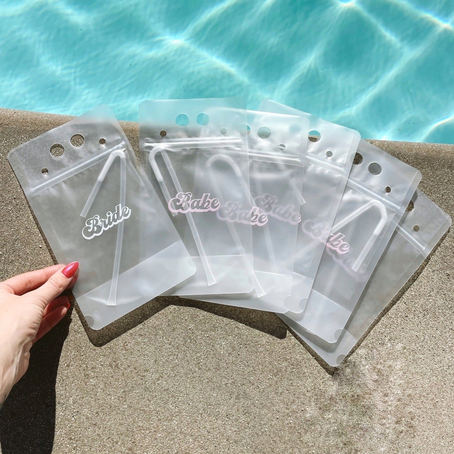 A woman&#39;s hand touching a transparent drink pouch that has cursive lettering on the front that reads &quot;Bride&quot; in white text. Next to that pouch are 5 identical pouches that say &quot;Babe&quot; in light pink text across the front. All 6 pouches are laying next to a pool of water.
