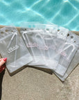 A woman's hand touching a transparent drink pouch that has cursive lettering on the front that reads "Bride" in white text. Next to that pouch are 5 identical pouches that say "Babe" in light pink text across the front. All 6 pouches are laying next to a pool of water.