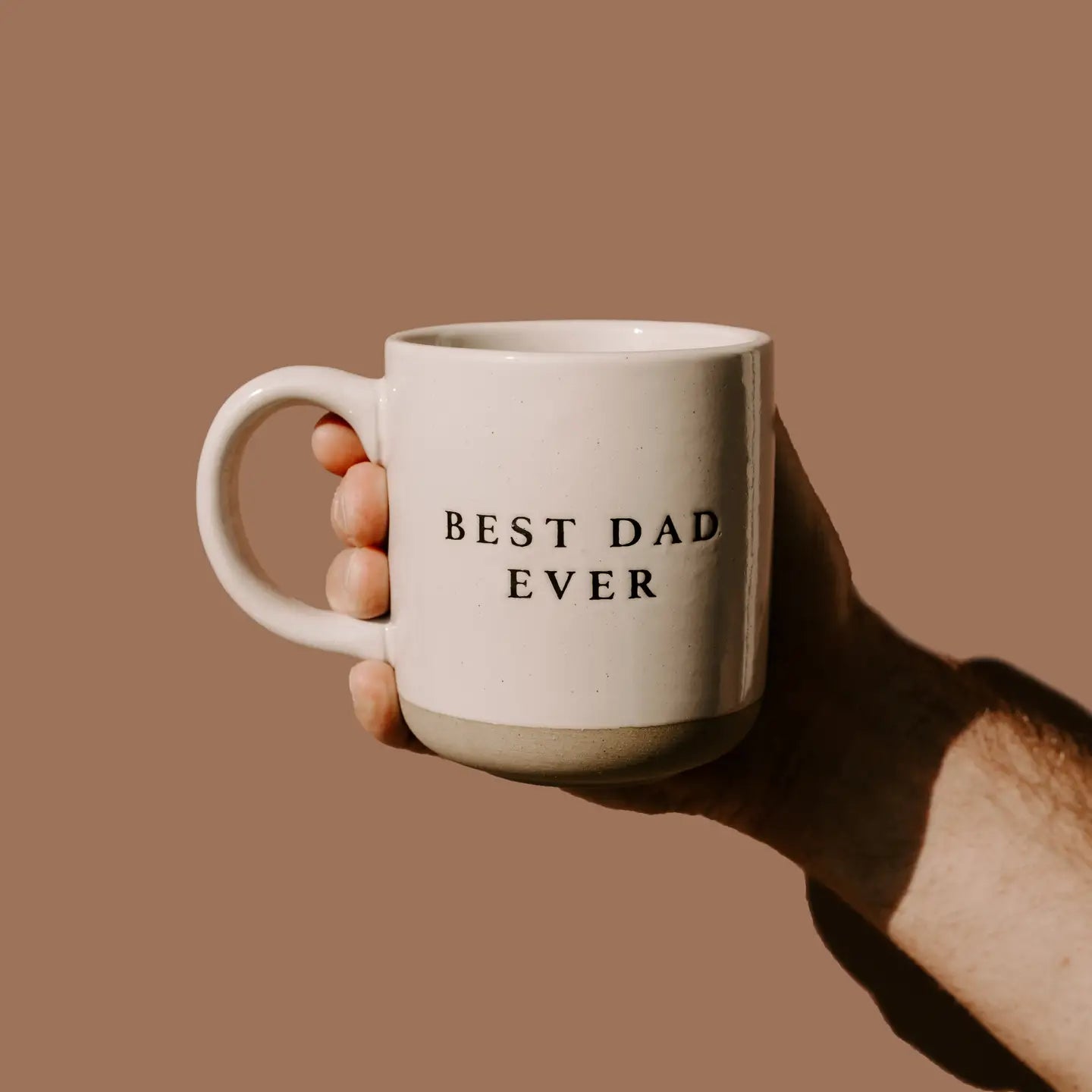 Hand holding a mug with 'Best Dad Ever' text on a brown background