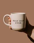 Hand holding a mug with 'Best Dad Ever' text on a brown background