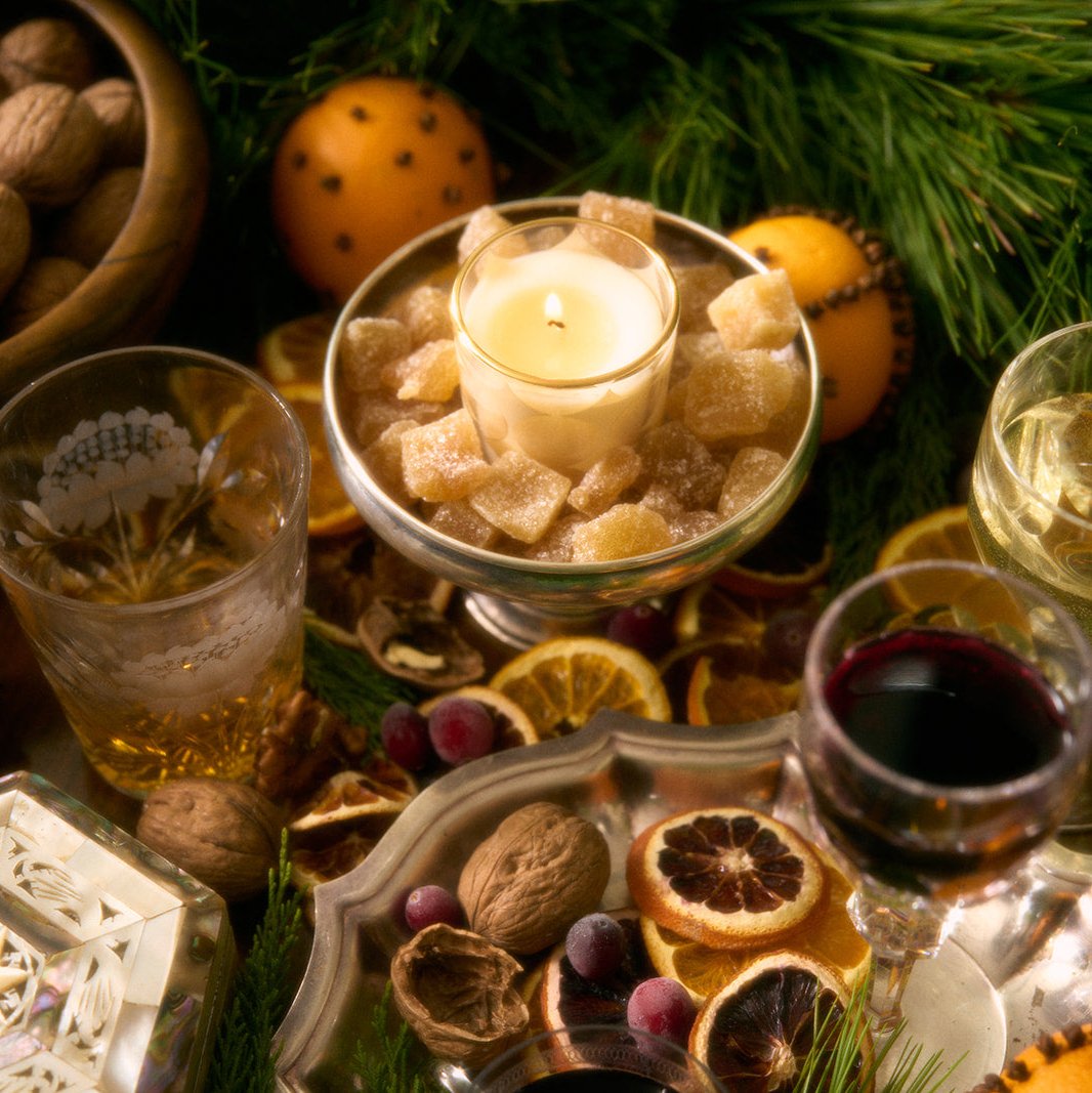 festive, holiday-themed arrangement. At the center is a candle in a glass votive, nestled among sugared ginger pieces on a silver pedestal dish. Surrounding centerpiece are elements of holiday decor, including whole walnuts, dried citrus slices, frosted cranberries, and sprigs of evergreen foliage. In the background, an orange studded with cloves a warm, aromatic touch. Various glasses filled with golden and red drinks suggest a celebratory setting, possibly mulled wine, whiskey. mood is cozy/elegant.