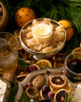 festive, holiday-themed arrangement. At the center is a candle in a glass votive, nestled among sugared ginger pieces on a silver pedestal dish. Surrounding centerpiece are elements of holiday decor, including whole walnuts, dried citrus slices, frosted cranberries, and sprigs of evergreen foliage. In the background, an orange studded with cloves a warm, aromatic touch. Various glasses filled with golden and red drinks suggest a celebratory setting, possibly mulled wine, whiskey. mood is cozy/elegant.