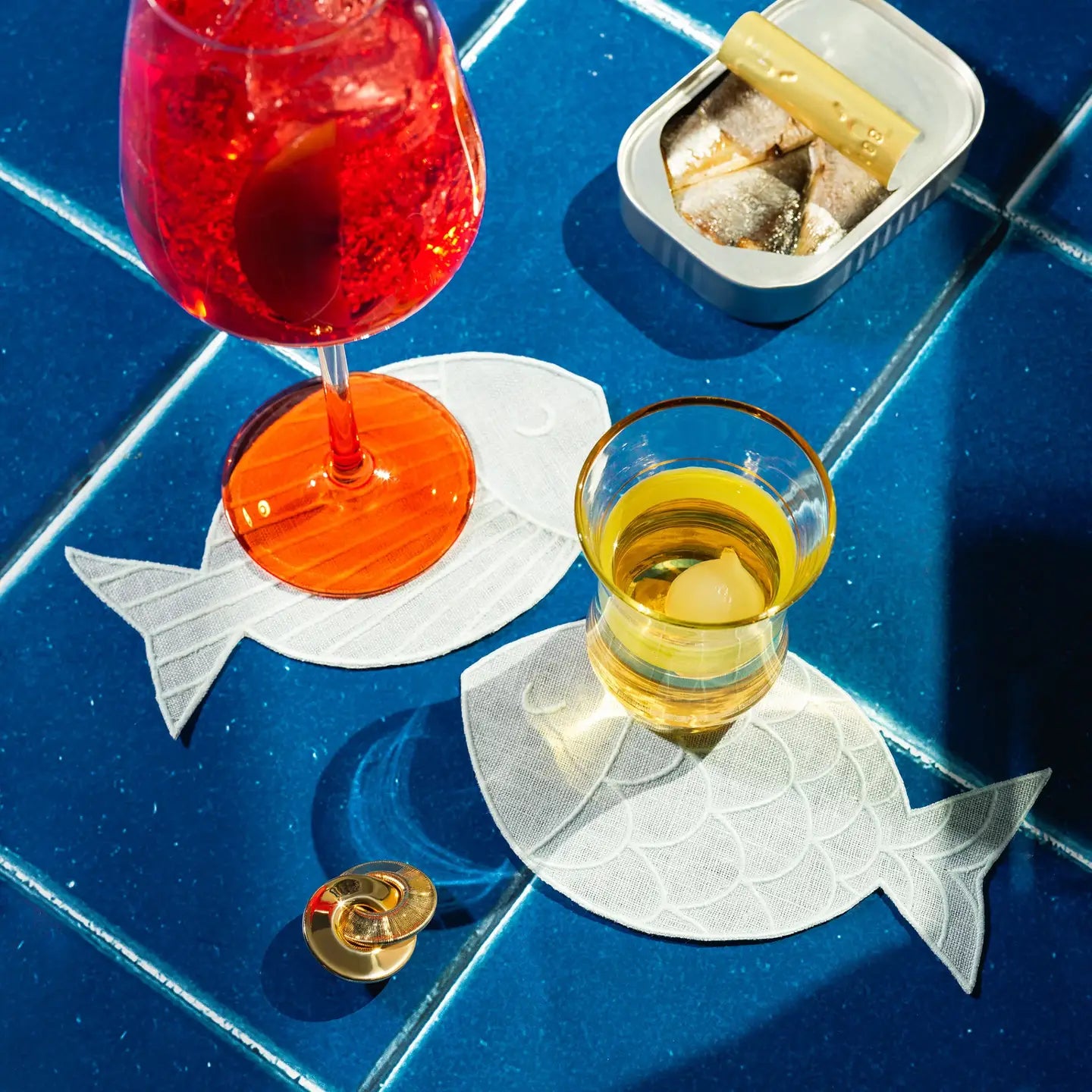 Two glasses of red and yellow drinks on fish-shaped coasters with a blue tiled background.