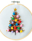 white fabric with black Christmas tree printed on it. Embroidered onto the tree are multicolored ornaments and a gold star tree topper. Embroidery hoop is light brown with silver metal clasp