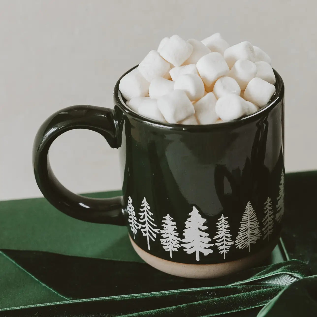 Black mug with white tree pattern filled with marshmallows on a green surface.