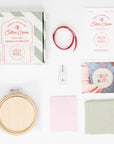 This image displays the contents of a "C'EST NOËL" cross-stitch kit by Cotton Clara. The kit includes a green-and-white striped box with the Cotton Clara logo, a wooden embroidery hoop, a piece of cross-stitch fabric (aida cloth), a needle, pink tissue paper, red ribbon, a hook for hanging, and printed instructions. There is also a reference card showing the finished cross-stitch hoop being held by someone, giving a glimpse of the final product. all materials presented on a clean white background. 