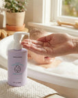 Person applying bubble bath to a hand with a bottle of bubble bath in a bathroom setting.