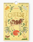 Illustrated cover of the 2025 Cheese Calendar by Rifle Paper Co. The design features a yellow background with a cow, cheeses, floral decorations, and text that reads 'CHEESE Please! Illustrated Calendar.