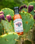 Bottle of Bushfire Farms Hickory Pear BBQ Sauce on a cactus plant with purple fruits.