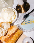 Butter dish with a stick of butter, bread, and a knife on a white surface.