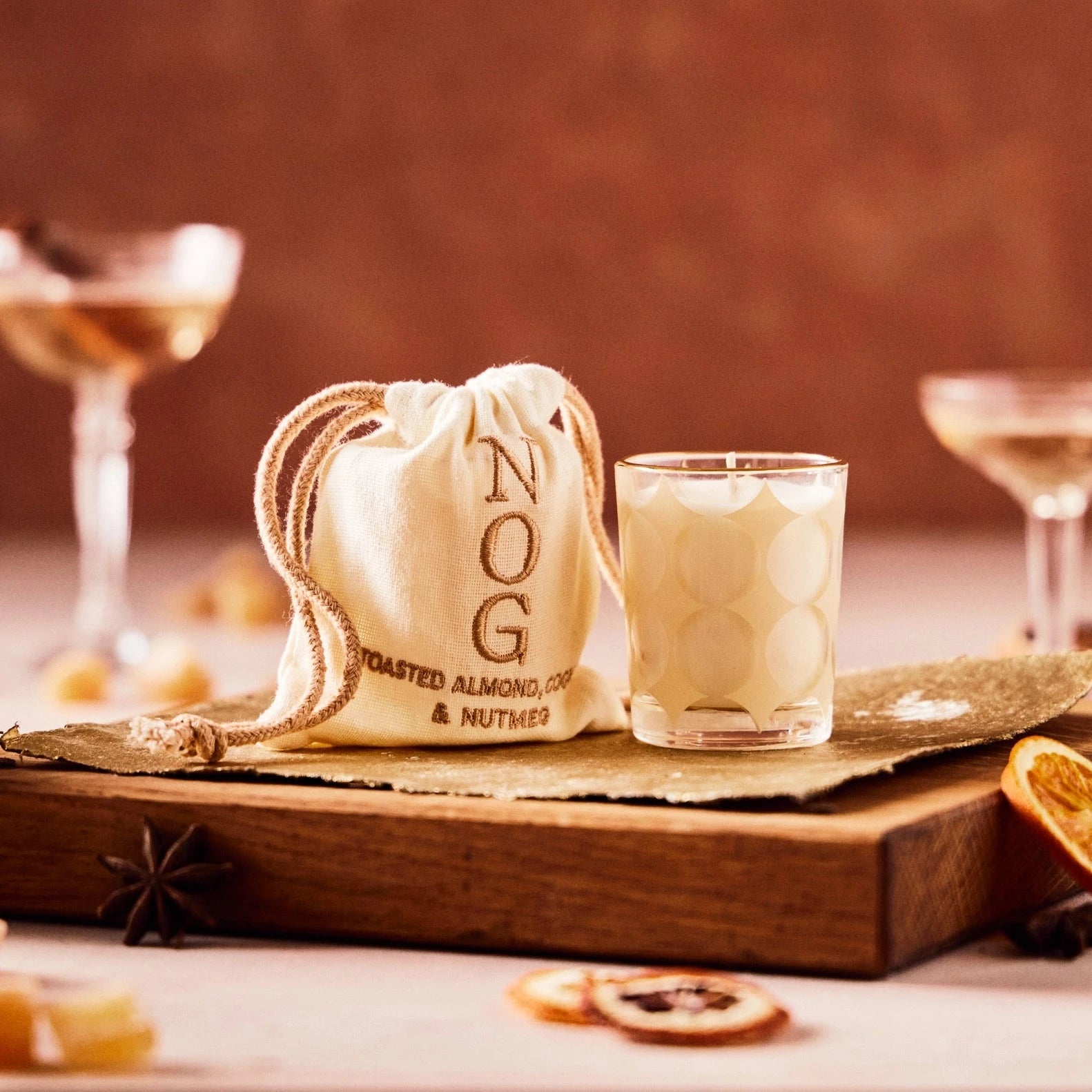 This image shows a cozy setup featuring a small cream-colored drawstring pouch labeled "NOG," with the additional text "Toasted Almond, Coco & Nutmeg" embroidered on it. Next to the pouch is a glass filled with a creamy beverage, possibly resembling eggnog or a similar festive drink. The backdrop is warm-toned, with hints of blurred drinks and garnishes, such as dried citrus slices and anise stars, adding to the cozy, seasonal atmosphere.