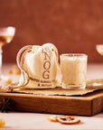 This image shows a cozy setup featuring a small cream-colored drawstring pouch labeled "NOG," with the additional text "Toasted Almond, Coco & Nutmeg" embroidered on it. Next to the pouch is a glass filled with a creamy beverage, possibly resembling eggnog or a similar festive drink. The backdrop is warm-toned, with hints of blurred drinks and garnishes, such as dried citrus slices and anise stars, adding to the cozy, seasonal atmosphere.