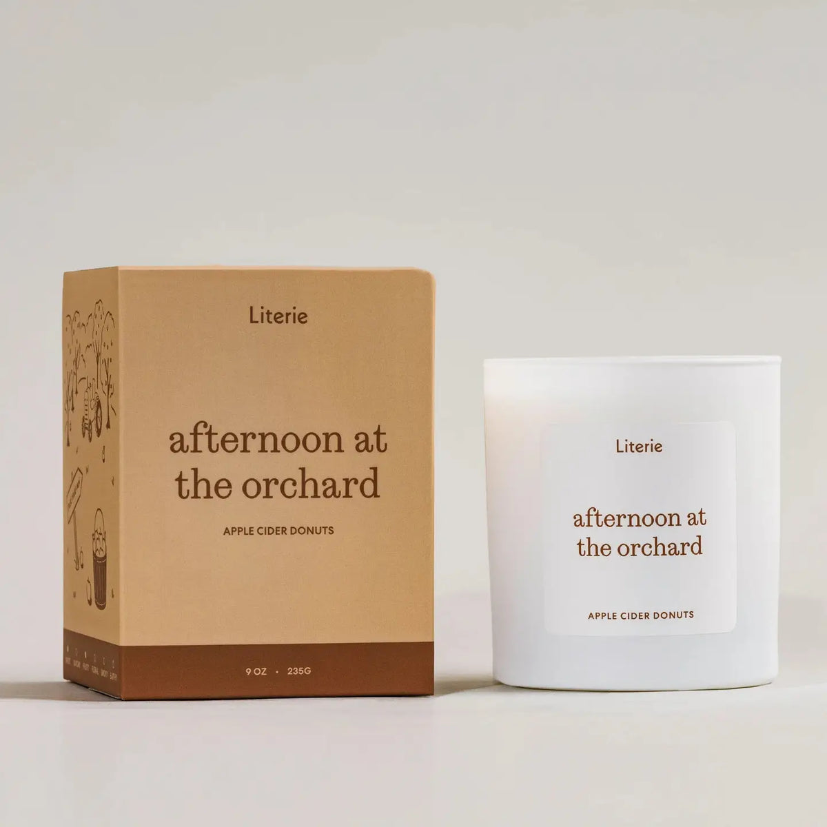 A white glass jar candle and its kraft paper box from the brand Literie, both labeled 'afternoon at the orchard' and 'Apple Cider Donuts', set against a neutral light gray background.