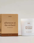 A white glass jar candle and its kraft paper box from the brand Literie, both labeled 'afternoon at the orchard' and 'Apple Cider Donuts', set against a neutral light gray background.