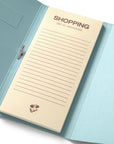 A light blue notebook with a cream-colored lined notepad insert that reads 'SHOPPING list & essentials,' photographed on a clean white background.
