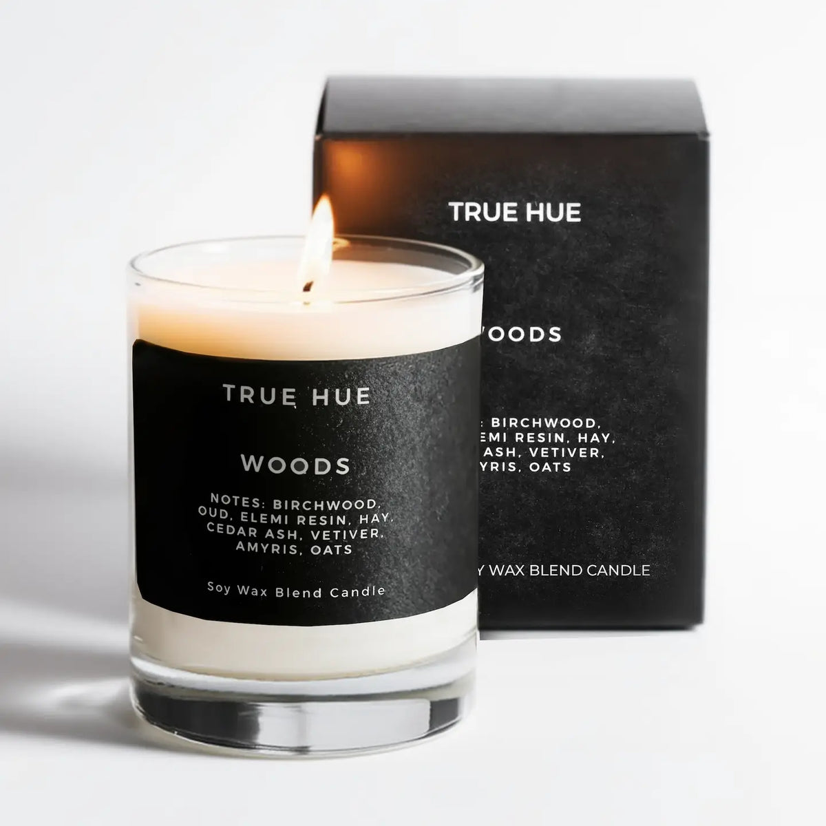A lit True Hue Woods soy wax candle in a clear glass jar with a black label, standing in front of its black branded box on a clean white background.