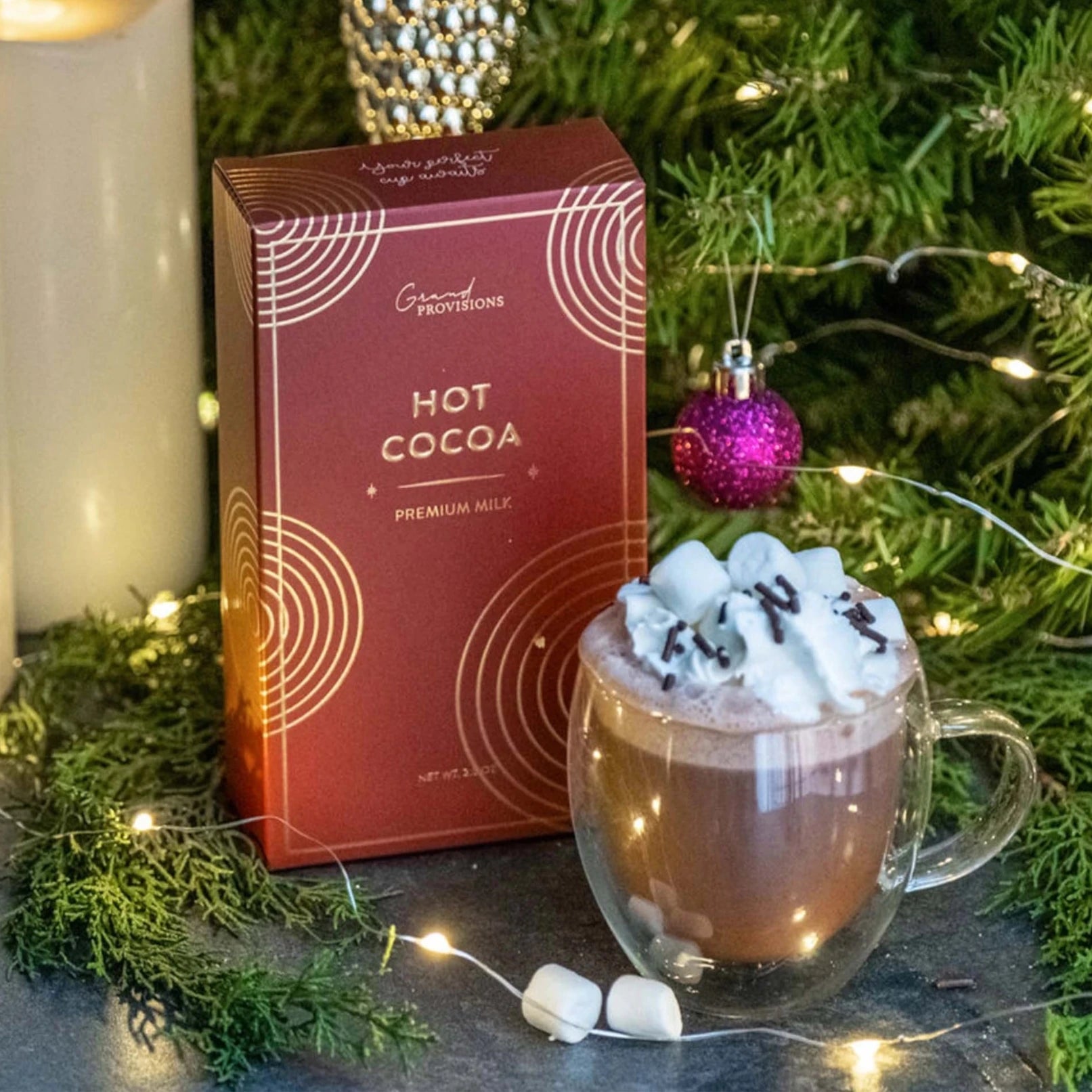 This image features a box of "Hot Cocoa" from Grand Provisions. The packaging is a rich, dark red with elegant, concentric circle patterns in gold, giving it a premium feel. The text "HOT COCOA" and "PREMIUM MILK" is printed in gold, emphasizing the high-quality product. The background is a simple, muted concrete texture, allowing the packaging to stand out.