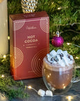 This image features a box of "Hot Cocoa" from Grand Provisions. The packaging is a rich, dark red with elegant, concentric circle patterns in gold, giving it a premium feel. The text "HOT COCOA" and "PREMIUM MILK" is printed in gold, emphasizing the high-quality product. The background is a simple, muted concrete texture, allowing the packaging to stand out.