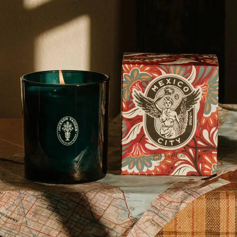 A dark teal-green glass jar candle next to its patterned red, orange, and teal box labeled 'Mexico City', styled on a map with warm sunlight and shadows.
