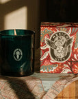 A dark teal-green glass jar candle next to its patterned red, orange, and teal box labeled 'Mexico City', styled on a map with warm sunlight and shadows.