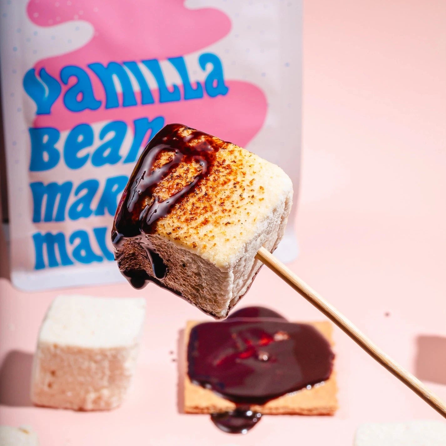 A close-up shot shows a perfectly toasted marshmallow from Mojave Mallows, coated with chocolate sauce, skewered on a stick. The package is visible in the background with other marshmallows and graham crackers, evoking a sense of indulgence and fun.