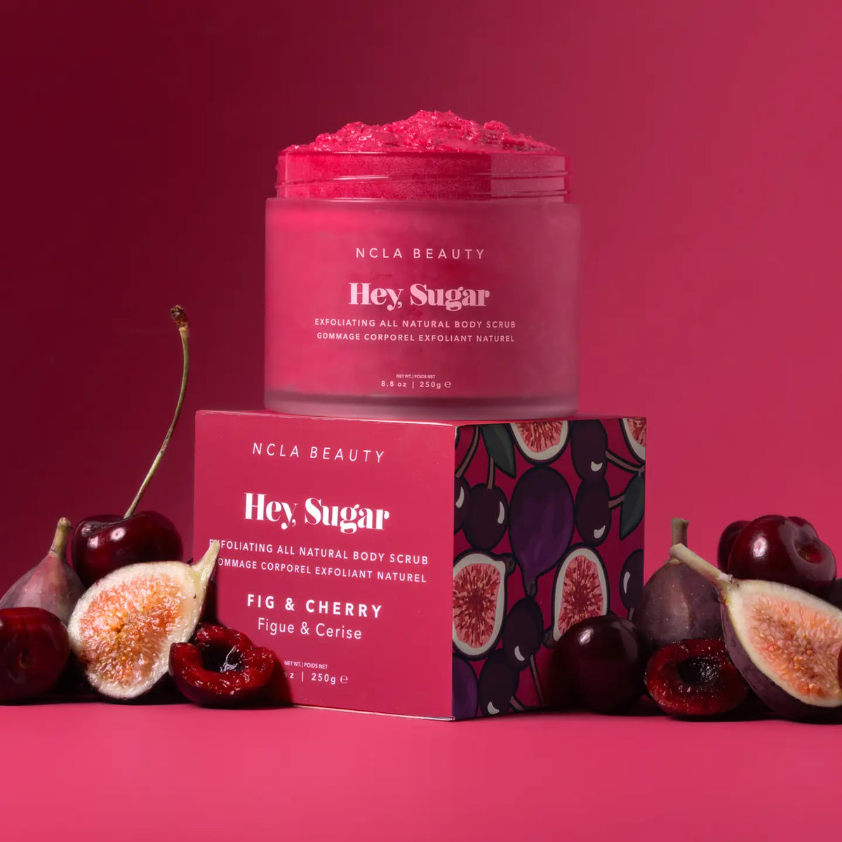 NCLA BEAUTY Hey Sugar exfoliating body scrub in a frosted jar, displayed on top of its pink packaging box with a fig and cherry pattern. The product is surrounded by fresh cherries and sliced figs on a solid pink background.