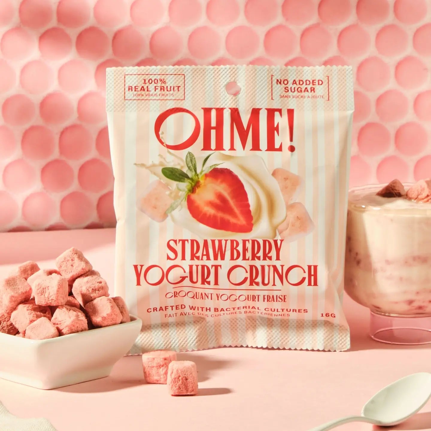 A bag of Ohme! Strawberry Yogurt Crunch is shown on a pink surface with a pink honeycomb backdrop. A bowl of pink yogurt cubes sits to the left, and a glass of pink yogurt with fruit sits to the right.