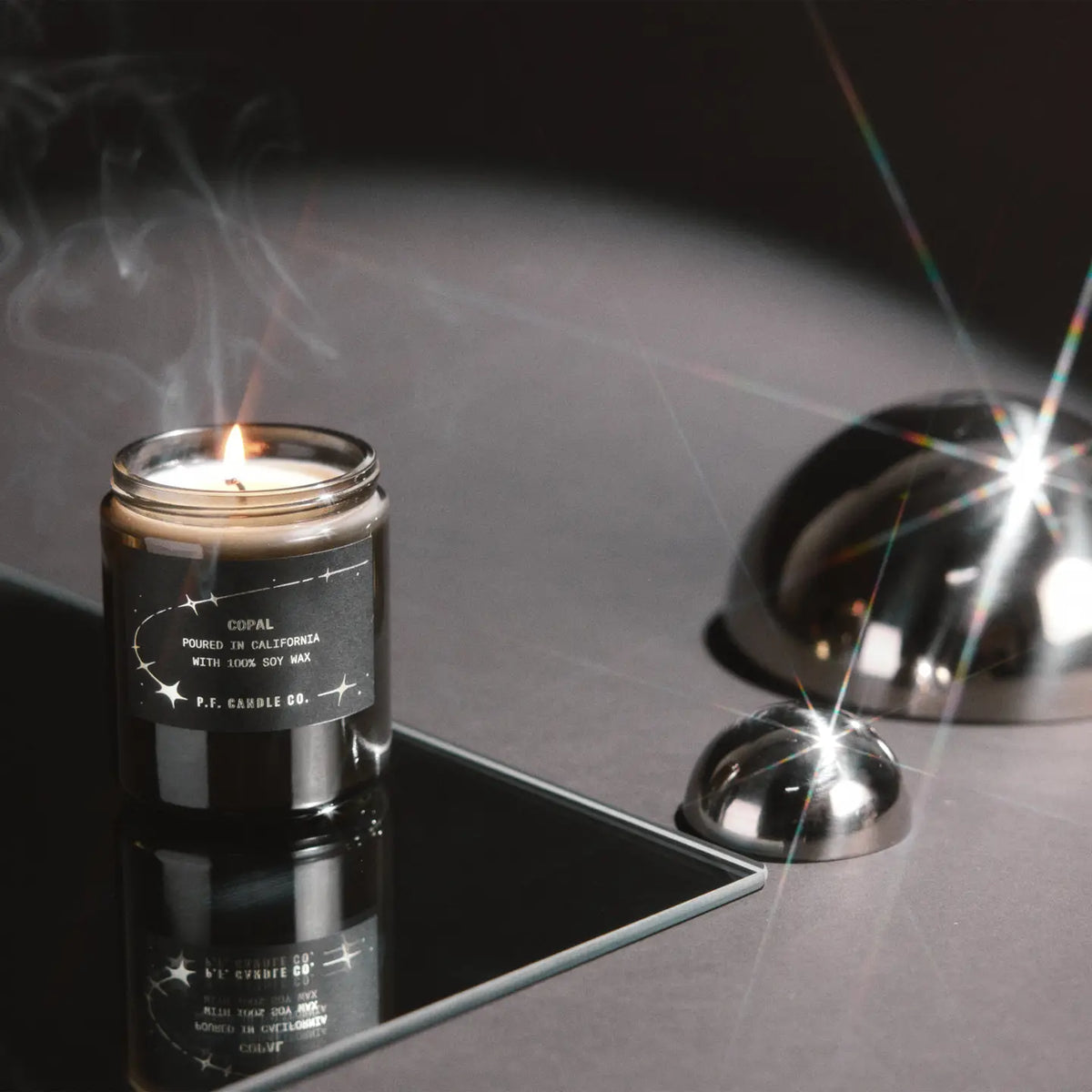 A lit candle in a dark glass jar sits on a reflective black surface, with the lid placed beside it and polished metal dome props reflecting light in a studio setup.