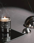A lit candle in a dark glass jar sits on a reflective black surface, with the lid placed beside it and polished metal dome props reflecting light in a studio setup.