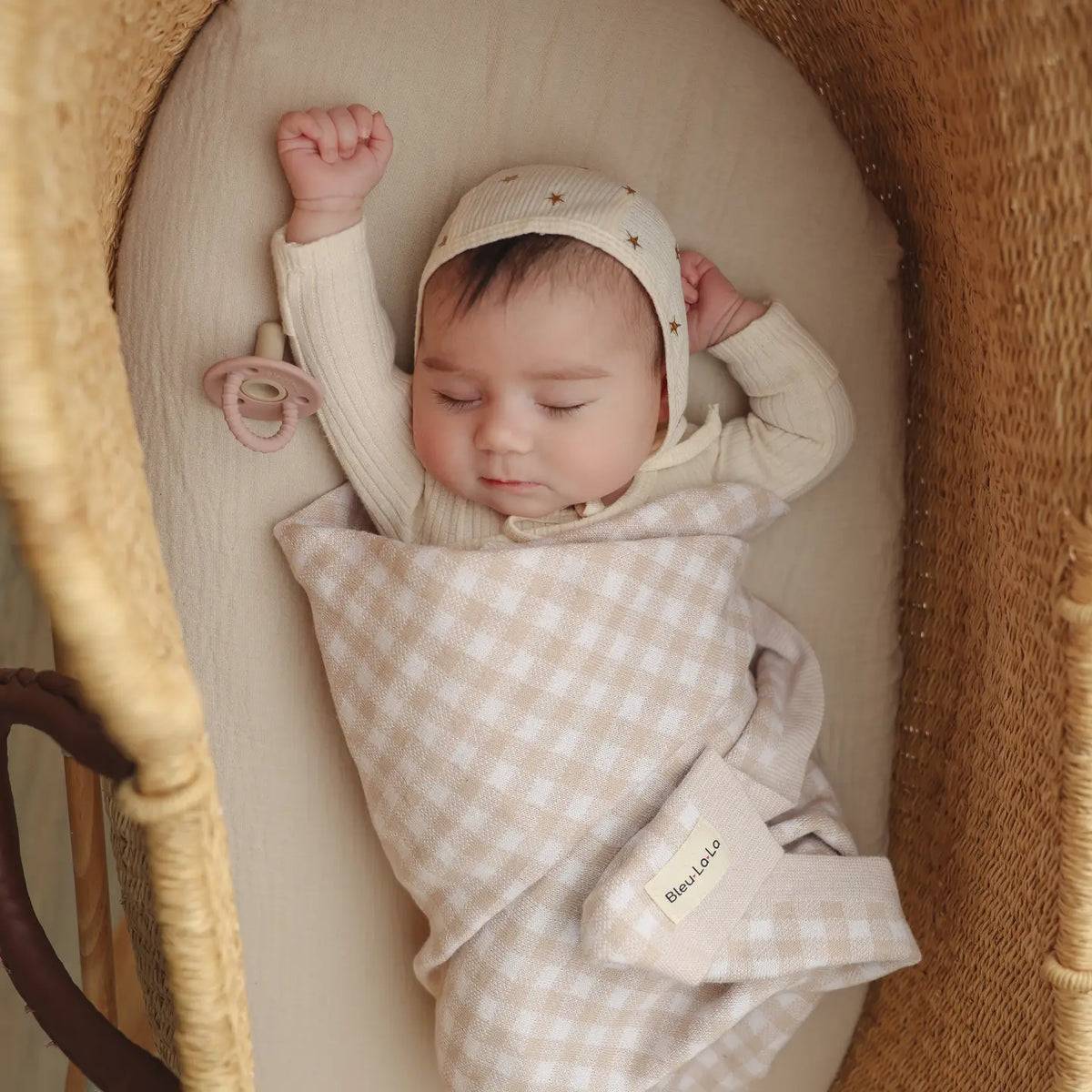 Baby sleeping in a moses basket with a checkered blanket and star-patterned hat.