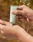 Hand holding a green skincare product with a blurred natural background