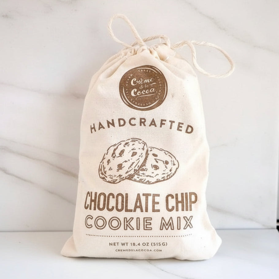 A bag of Crème Cocoa Handcrafted Chocolate Chip Cookie Mix stands on a white surface next to a bottle of vanilla bean syrup, with several chocolate chip cookies, white sugar, and dark chocolate pieces in front.