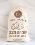 A bag of Crème Cocoa Handcrafted Chocolate Chip Cookie Mix stands on a white surface next to a bottle of vanilla bean syrup, with several chocolate chip cookies, white sugar, and dark chocolate pieces in front.