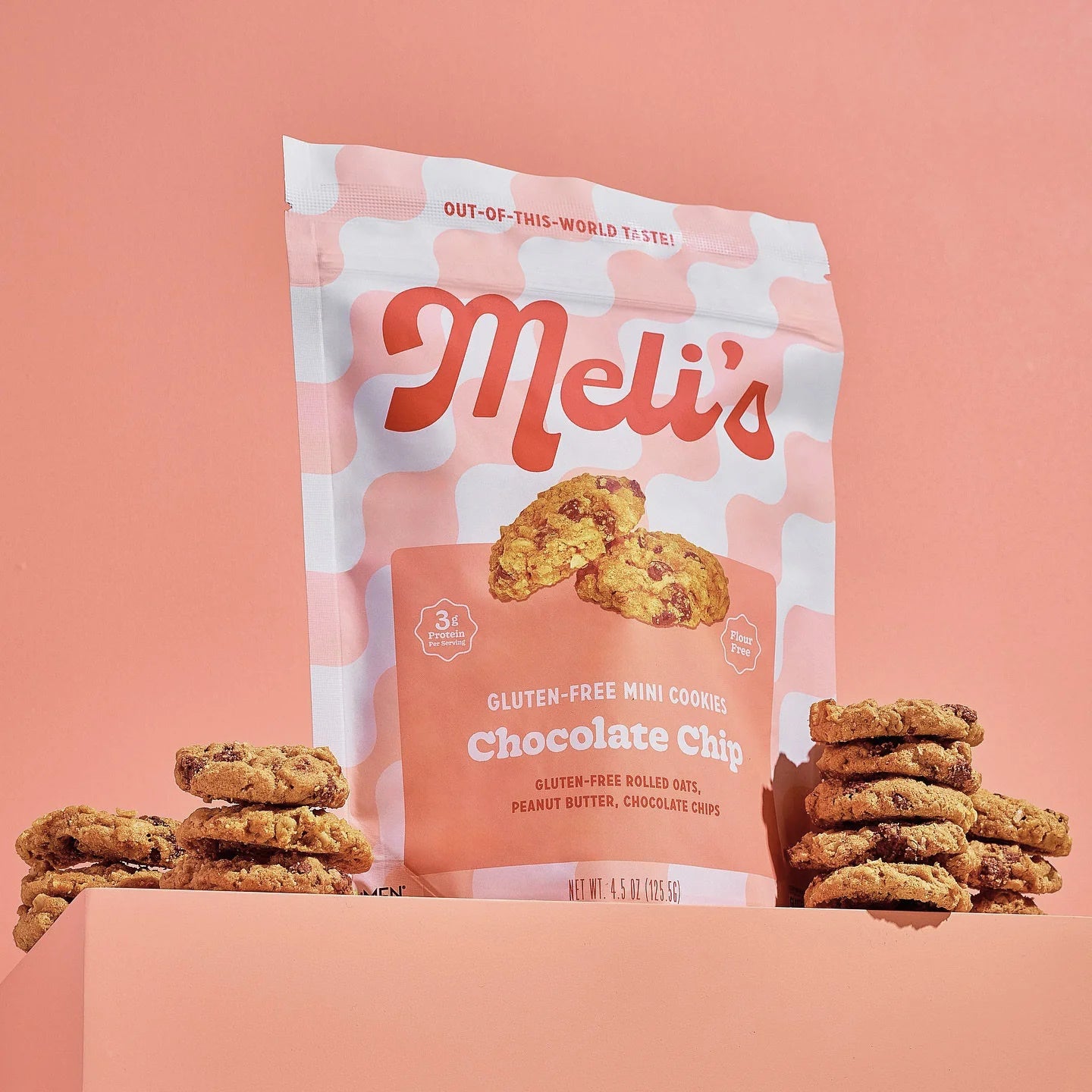 A stand-up pouch of Meli's Gluten-Free Mini Cookies, Chocolate Chip flavor, styled with stacks of cookies on either side against a solid peach-pink background.