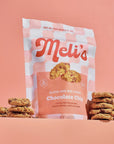 A stand-up pouch of Meli's Gluten-Free Mini Cookies, Chocolate Chip flavor, styled with stacks of cookies on either side against a solid peach-pink background.