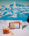 Toddy Kit with a glass of liquid in front of a mountainous landscape